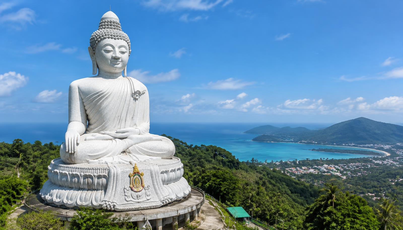 Large white seated Buddha statue on a green hilltop in Phuket overlooking a turquoise bay