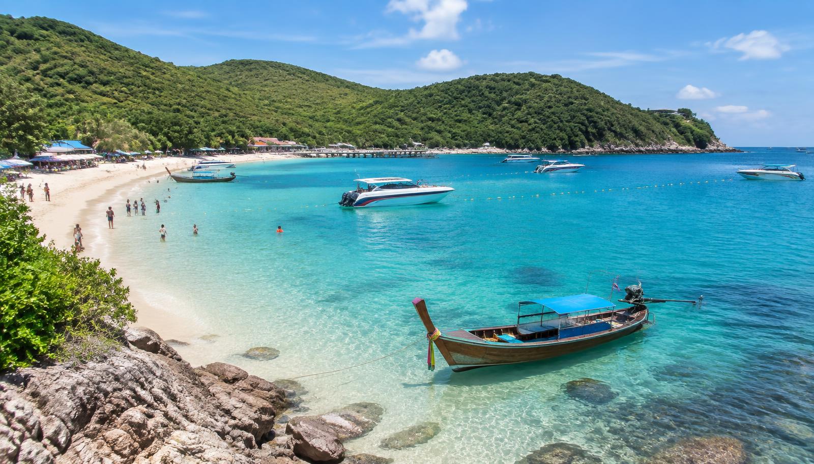 Clear turquoise water, a traditional longtail boat, and a sandy beach framed by green hills on Koh Larn island near Pattaya
