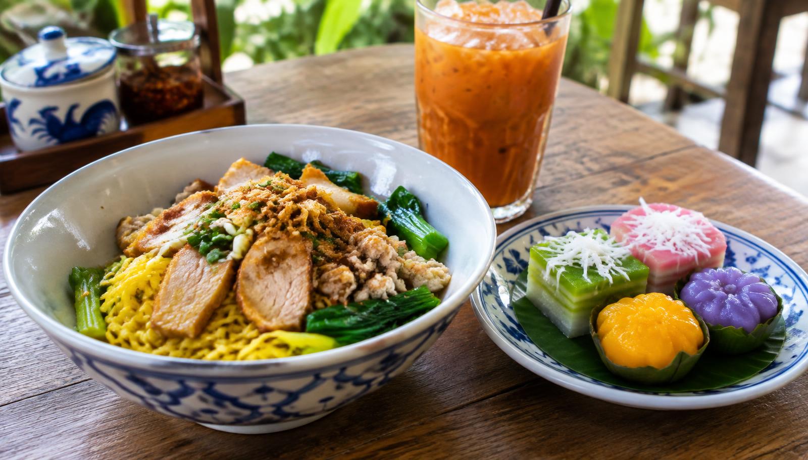 Bowl of yellow noodles with pork and greens beside colorful traditional desserts and an iced tea in Phuket