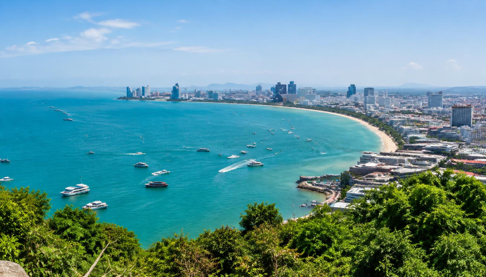 Panoramic view of Pattaya Bay with turquoise water, boats, and the city skyline along the curved beachfront, seen from a green hillside