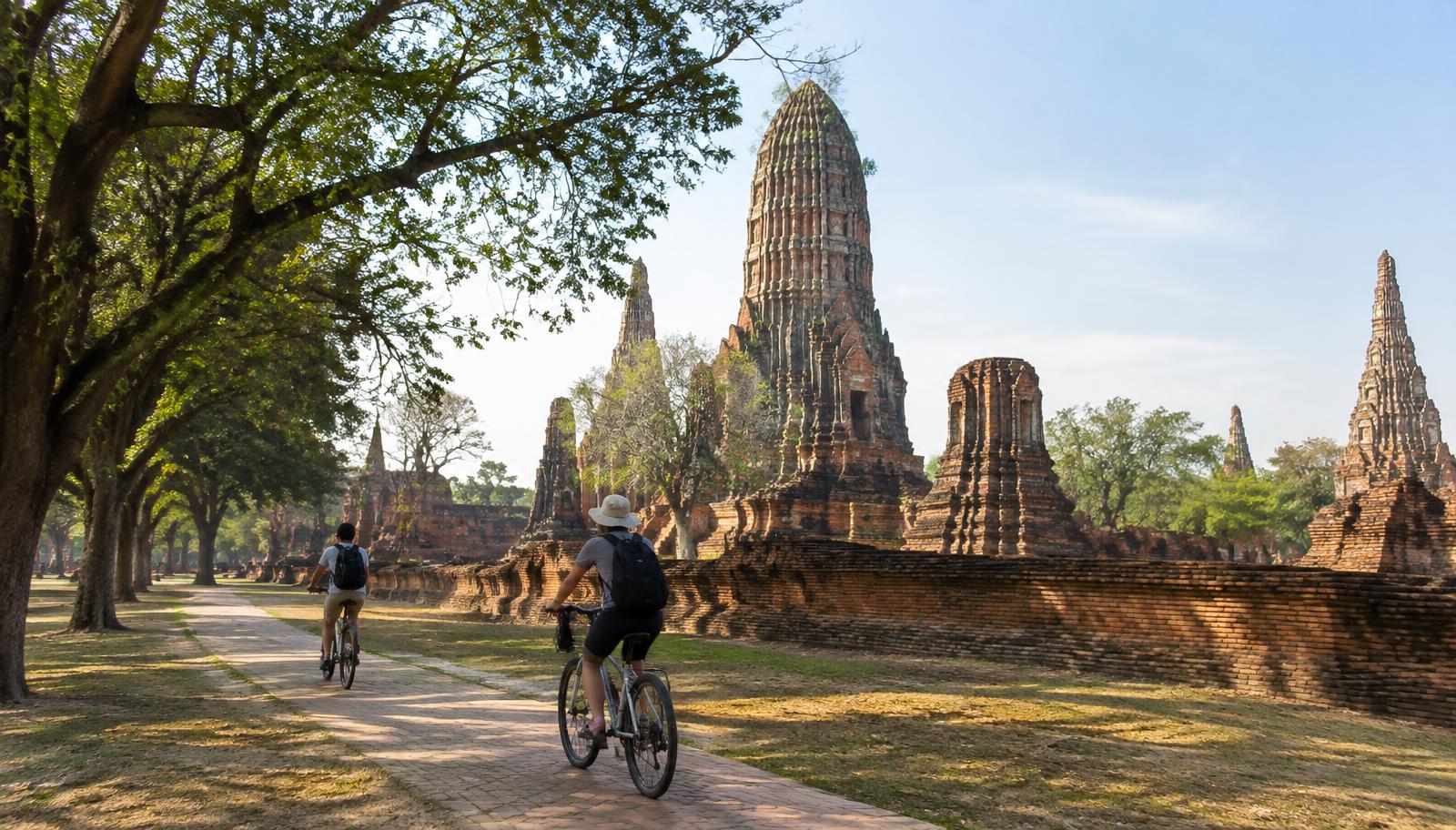 Travelers cycling on a shaded path past brick temple ruins in Ayutthaya Historical Park