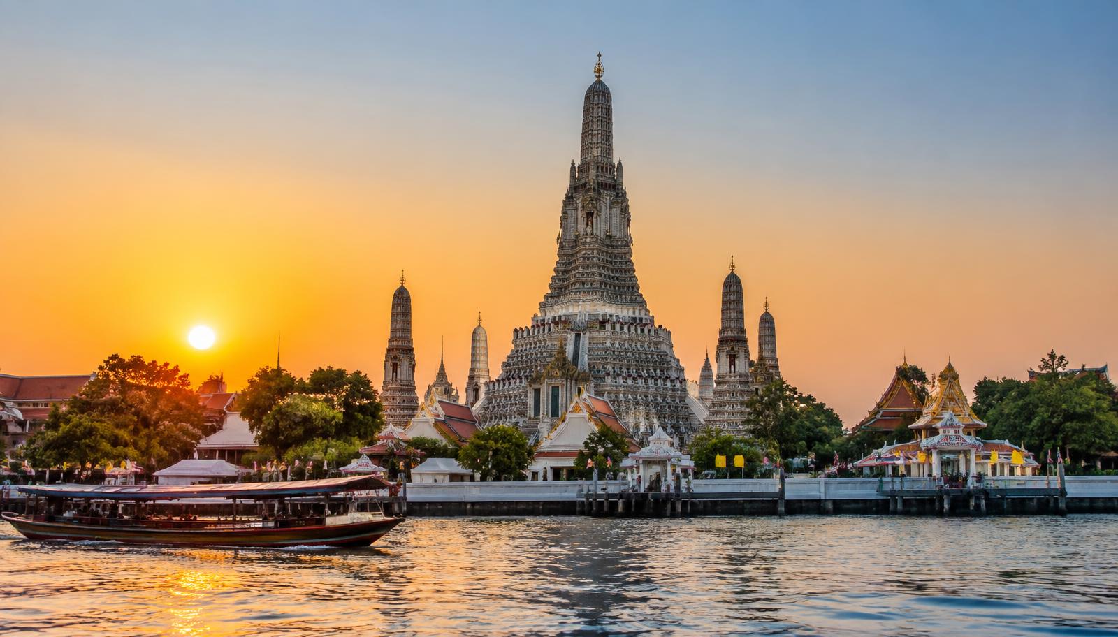 Wat Arun temple and its ornate spires beside the Chao Phraya River at sunset, with a long-tail boat passing
