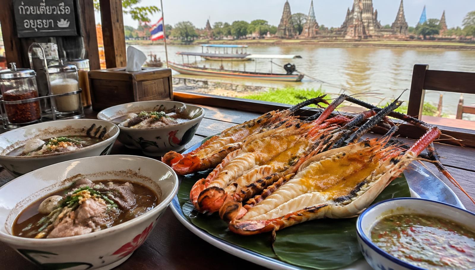 Bowls of boat noodles and grilled river prawns at a riverside restaurant with temple ruins behind
